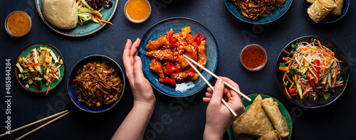 Set of Chinese dishes on table, female hands holding chopsticks: sweet and sour chicken, fried spring rolls, noodles, rice, steamed buns with bbq glazed pork, Asian style banquet or buffet, top view 