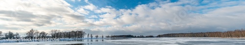 Beautiful snowy landscape on the river along the forest. Panorama of the coastal line in winter. Horizontal panorama of growing trees on the lake shore and clouds on a sunny winter day.