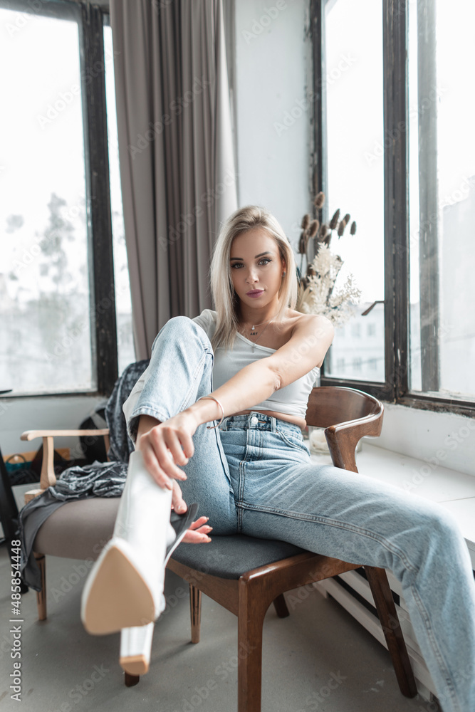 Cool fashion glamour blonde woman model in stylish clothes with blue vintage jeans put on white shoes in room. Trendy women’s leather footwear Cool fashion glamour blonde woman model in stylish clothes with blue vintage jeans put on white shoes in room. Trendy women’s leather footwear