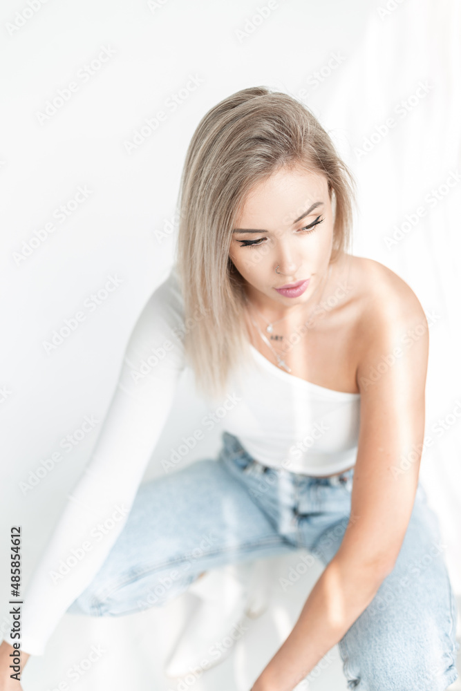 Delicate beautiful young blonde woman with a white t-shirt and vintage blue jeans sits on a white background in sunlight Delicate beautiful young blonde woman with a white t-shirt and vintage blue jeans sits on a white background in sunlight