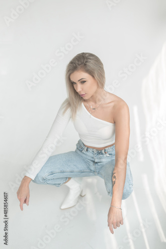 Lovely tender beautiful young blonde woman in a fashionable white T-shirt with blue jeans and leather shoes sits on a white background in the studio at sunlight