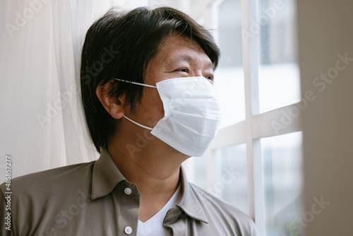 Lonely Korean man in medical facial mask looking through the window. solation at home for self quarantine. Unhappy male in facemask suffer from coronavirus. Quarantine, corona pandemic concept.