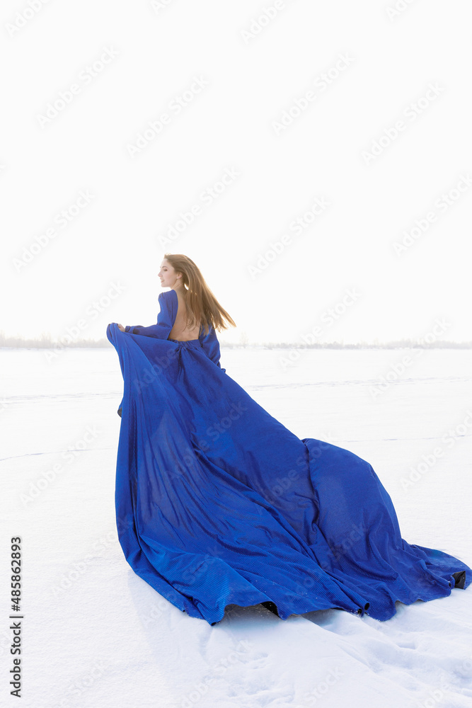 Fotka „Incredible stunning girl in a blue airy dress with a long train  running on the ice. In the background winter landscape frozen river covered  with ice. The woman in the dress