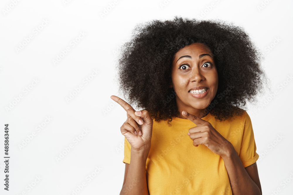 Close up portrait of awkward woman smiles, points at upper left corner empty space, copyspace for advertisement, stands in yellow tshirt over white background Close up portrait of awkward woman smiles, points at upper left corner empty space, copyspace for advertisement, stands in yellow tshirt over white background