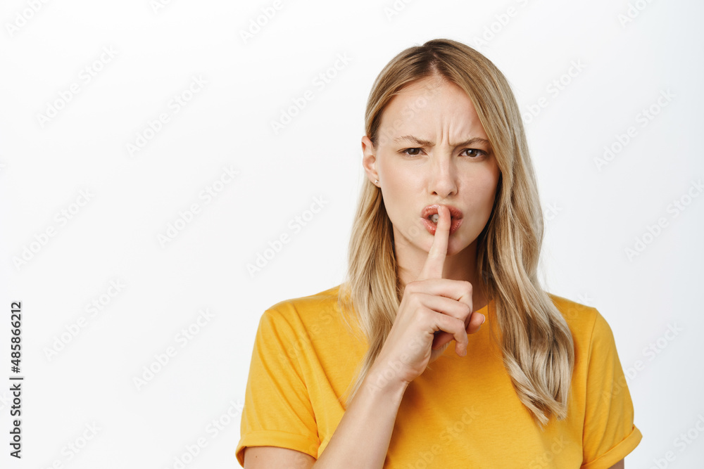 Portrait of angry blond girl shushing, hush with upset frowning face, making be quiet, silence gesture, standing in tshirt over white background Portrait of angry blond girl shushing, hush with upset frowning face, making be quiet, silence gesture, standing in tshirt over white background