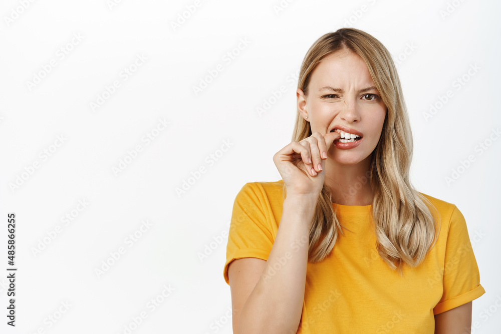 Nervous blond girl, anxious young woman biting fingernails, looking guilty or worried, standing against white background Nervous blond girl, anxious young woman biting fingernails, looking guilty or worried, standing against white background