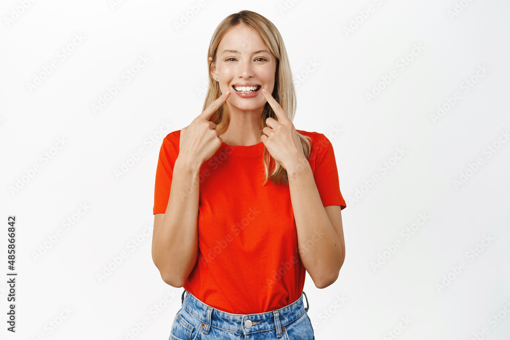 Portrait of smiling blond girl pointing at her white teeth, perfect smile, dental clinic advertisement, standing happy and pleased in red t-shirt, white background in studio Portrait of smiling blond girl pointing at her white teeth, perfect smile, dental clinic advertisement, standing happy and pleased in red t-shirt, white background in studio