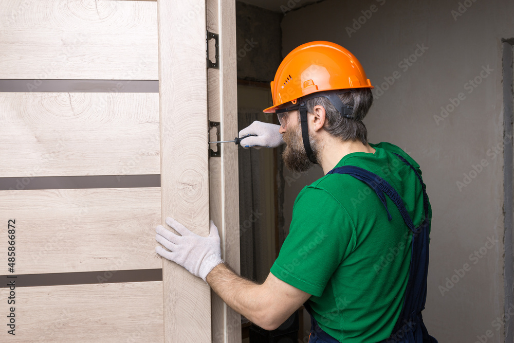 A male locksmith installs a door in an apartment. The guy works with a screwdriver. - obrazy, fototapety, plakaty 