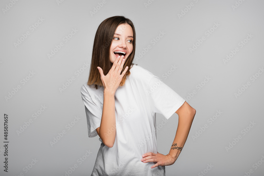laughing woman covering mouth with hand while standing with hand on hip isolated on grey laughing woman covering mouth with hand while standing with hand on hip isolated on grey