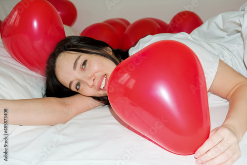 The smiling brunette woke up on a white bed with red balloons in the shape of a heart. A surprise gift for your beloved girl on Valentine's Day.