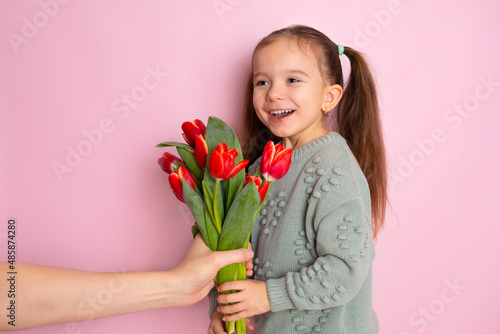 A little funny cute girl is given a bouquet of tulips and she reacts surprised. Pink background. Happy women's day. Space for text. Bright Emotions.