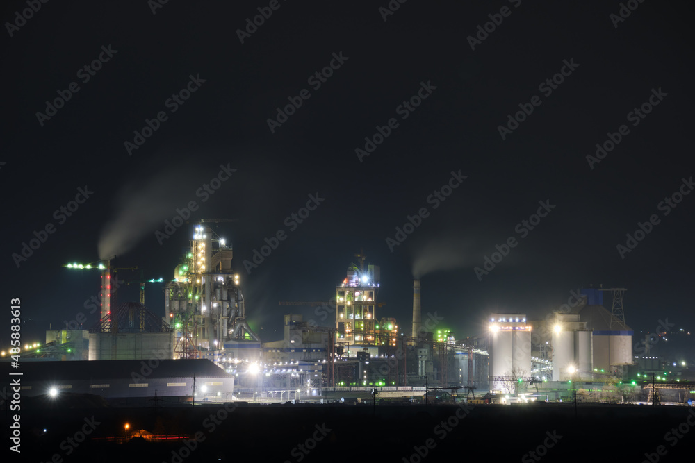 Illuminated cement plant with high factory structure and tower cranes at industrial production area at night. Manufacture and global industry concept