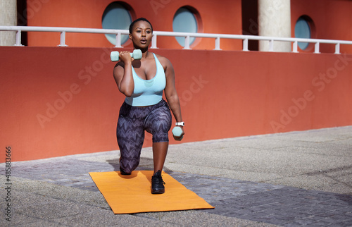 Your only worthy competitor is YOU. Shot of a young woman on a gym mat using dummbells against an urban background. photo