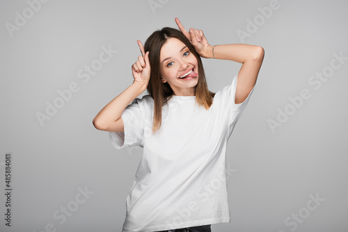 cheerful and tricky woman sticking out tongue and showing horns gesture isolated on grey