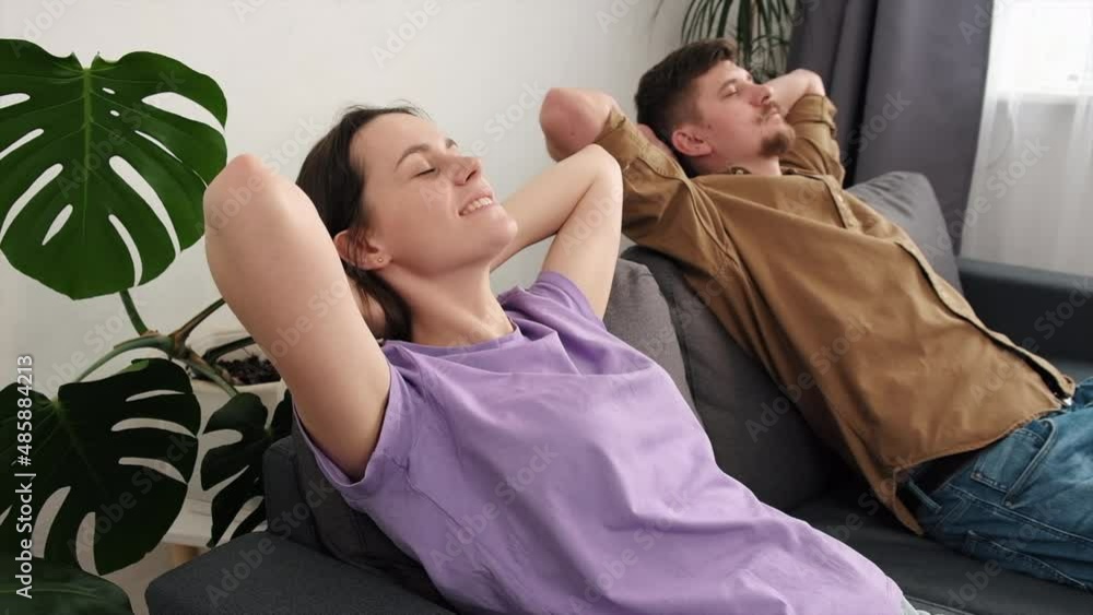 Close up of smiling husband and wife breathing fresh air, no stress in living room holding hands behind head. Calm serene lazy young couple relaxing sitting on cozy grey sofa at home with eyes shut Close up of smiling husband and wife breathing fresh air, no stress in living room holding hands behind head. Calm serene lazy young couple relaxing sitting on cozy grey sofa at home with eyes shut