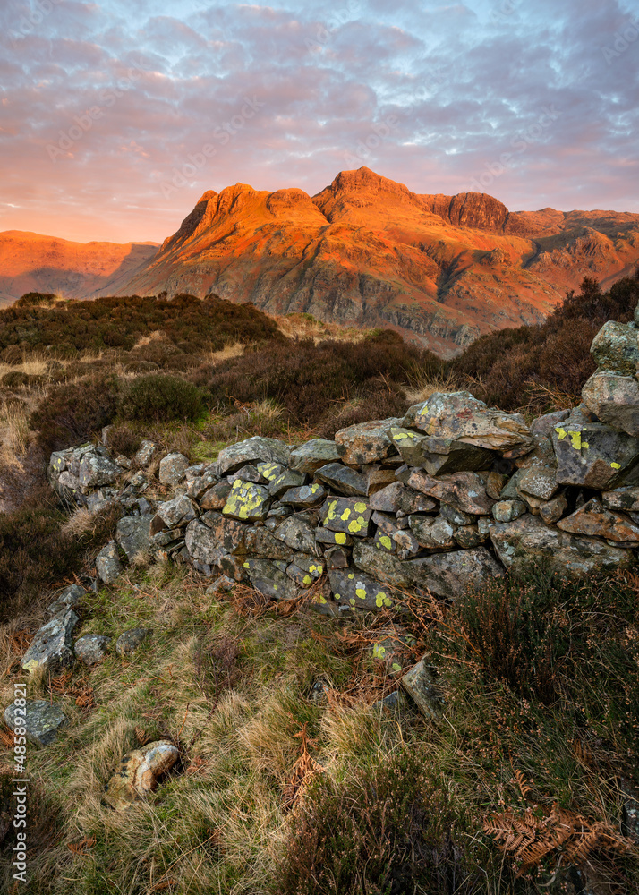 Golden light at sunrise on Langdale Pikes mountain peaks with old stone wall in rural landscape. Lake District National Park, UK.