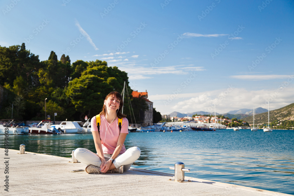 Smiling young woman sitting cross-legged on pier, listening to music with Croatian coastline in background. Bright sunny day in Dubrovnik, Croatia. Travel lifestyle concept