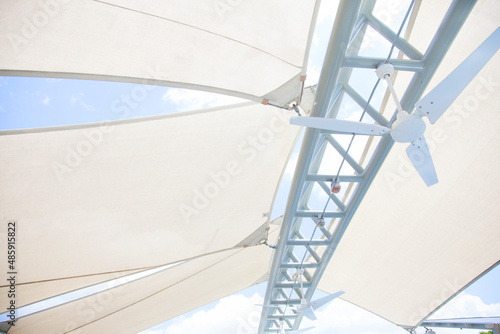 Looking up at a bright white outdoor canvas canopy and geometric architectural strut with a ceiling fan, protecting patio from the summer sun and bright blue sky photo