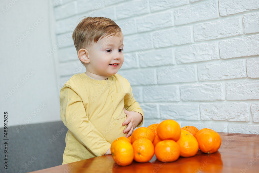 A little boy 2 years old is standing at a table with tangerines. Baby wants to sit down citrus fruits for the first time