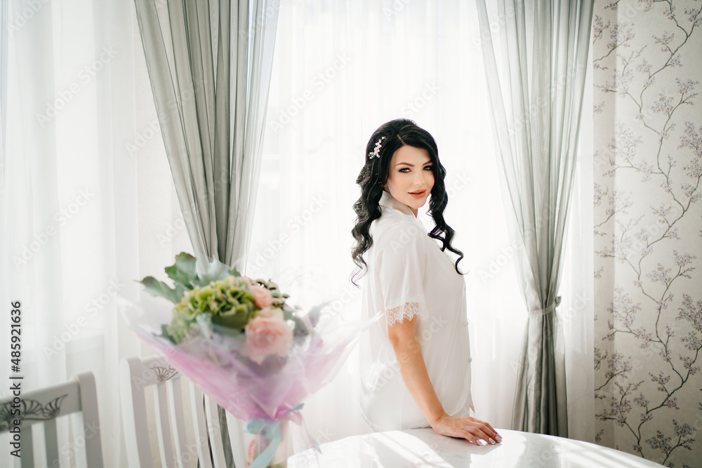 pregnant bride brunette in white pajamas by window and table with bouquet. pregnant bride brunette in white pajamas by window and table with bouquet.