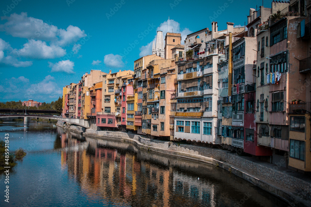 Panorama of Girona from the river, looking away from Girona cathedral of Saint Mary and red Eiffel bridge. Greenery on the water is seen.