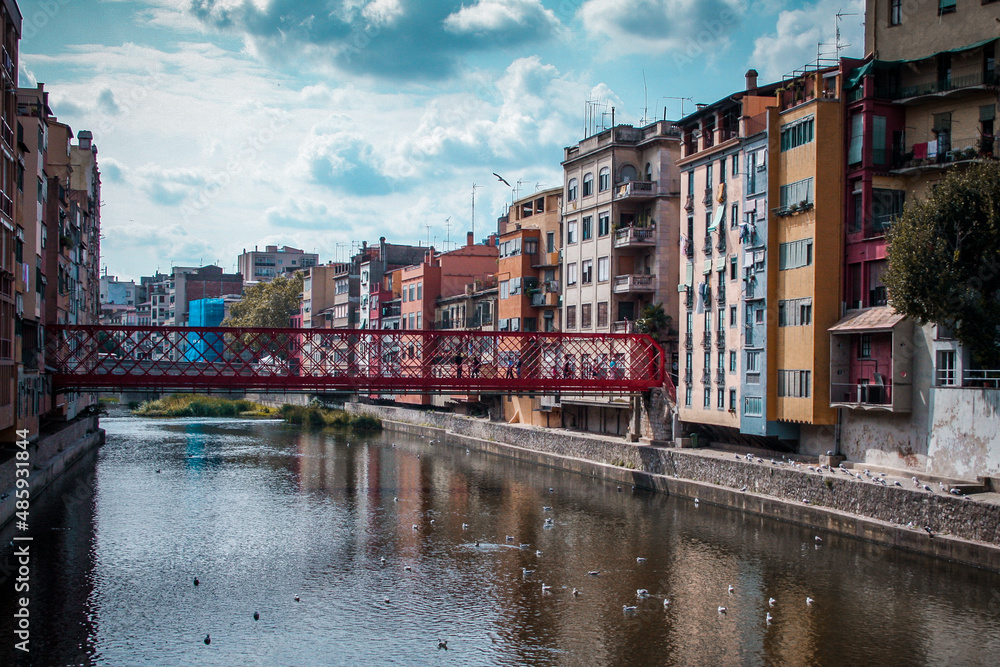 Panorama of Girona from the river, looking towards Girona cathedral of Saint Mary and red Eiffel bridge. Greenery on the water is seen.