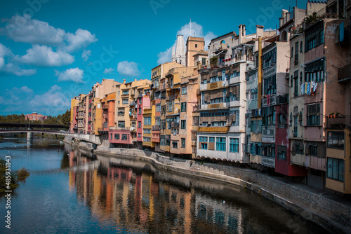 Panorama of Girona from the river, looking away from Girona cathedral of Saint Mary and red Eiffel bridge. Greenery on the water is seen.