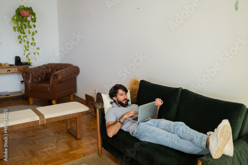 Young business man working at home from his living room couch with laptop on his lap. Man lying down. Home office concept.