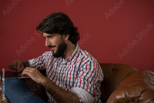 Young business man working at home with laptop on a brown arm chair with a red painted wall in the background. Home office concept.