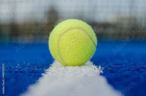 Selective focus. Ground level view of a ball on the line of a blue paddle tennis court with the net out of focus in the background. © Vic