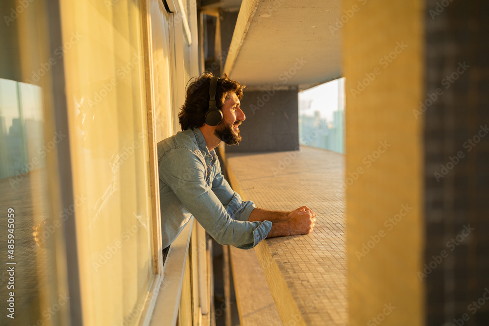 Side view of young man listening to music with headphones in his ears by the window with a city view landscape in the background. High quality photo