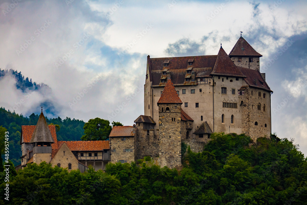 Stone fortified Gutenberg Castle among green trees on top of hill on background with mountains and cloudy sky, Balzers, Liechtenstein