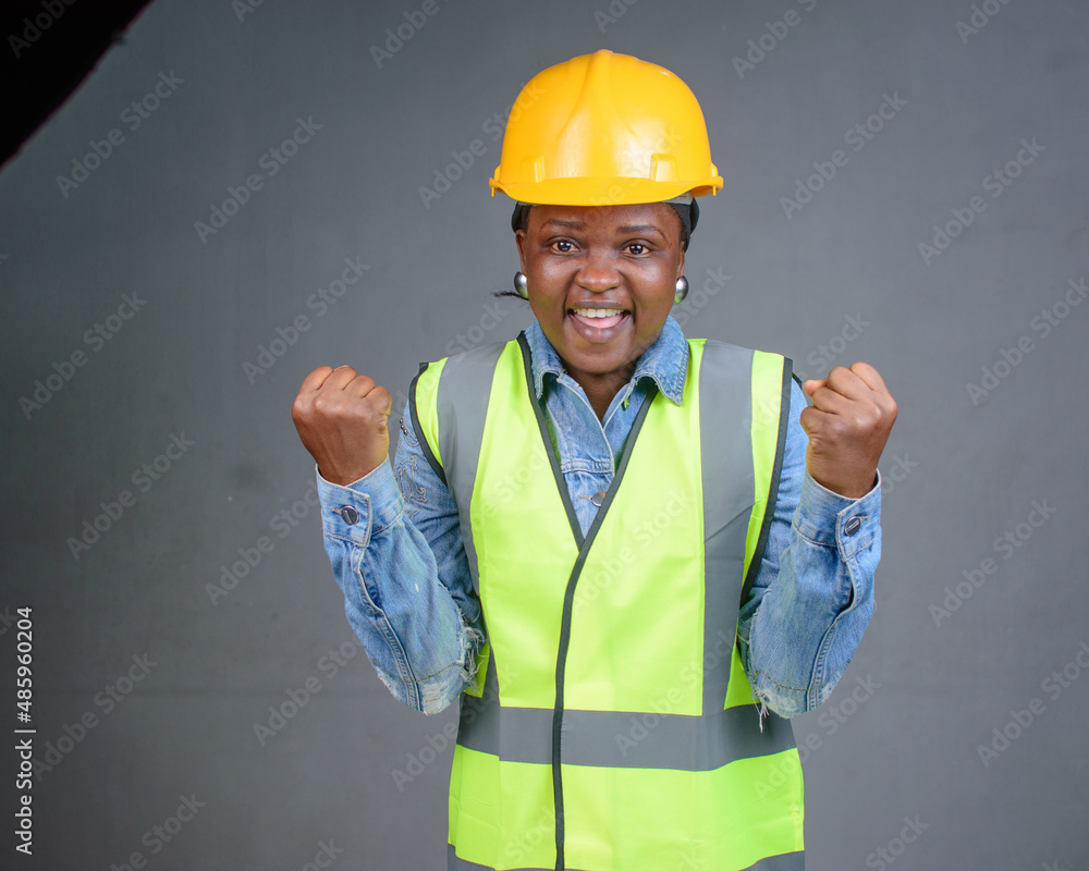 Studio portrait of a happy African Nigerian career lady or female engineer wearing a yellow safety helmet and a reflective jacket and raising her fits up in jubilation Studio portrait of a happy African Nigerian career lady or female engineer wearing a yellow safety helmet and a reflective jacket and raising her fits up in jubilation