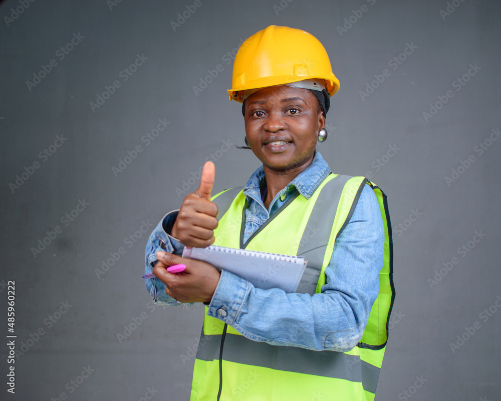 Studio portrait of a happy African Nigerian career lady or female engineer wearing a yellow safety helmet, reflective jacket and having a pen and writing pad in her hands while doing thumbs up