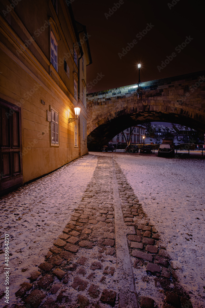 snow paved sidewalk and light from street lights next to Charles Bridge in the center of Prague in winter