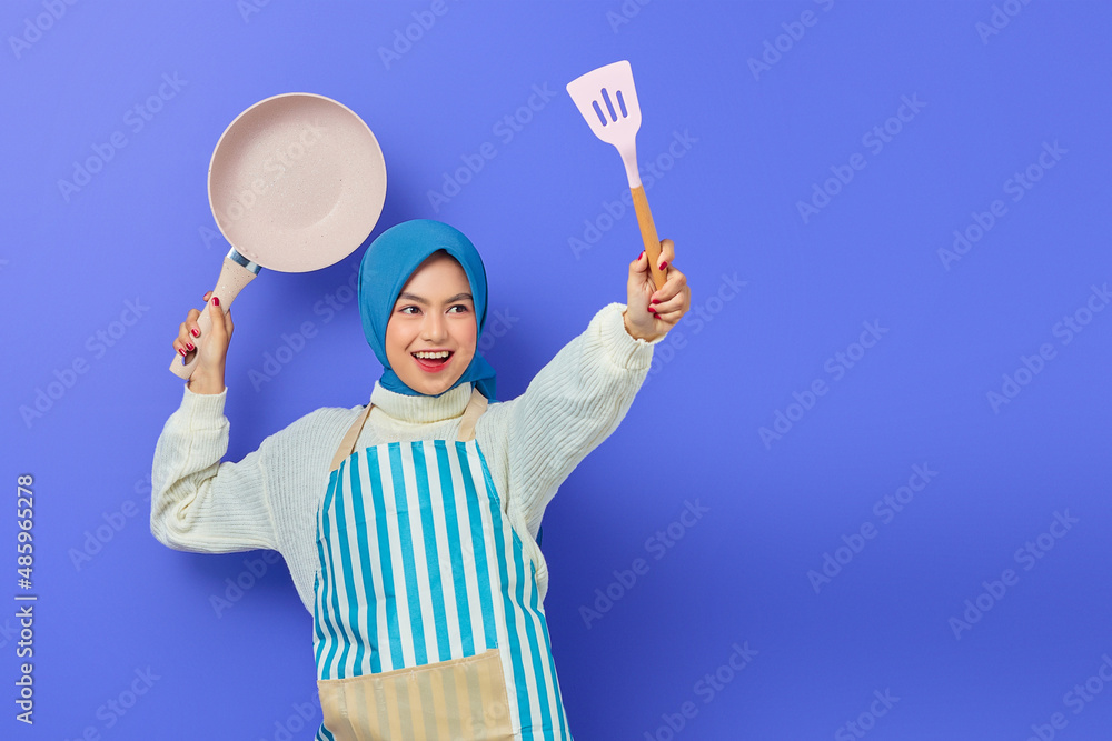 Cheerful beautiful Asian woman in white sweater covered in apron and hijab hold spatula and pan attack enemies while doing housework isolated over purple background. Housekeeping concept Cheerful beautiful Asian woman in white sweater covered in apron and hijab hold spatula and pan attack enemies while doing housework isolated over purple background. Housekeeping concept