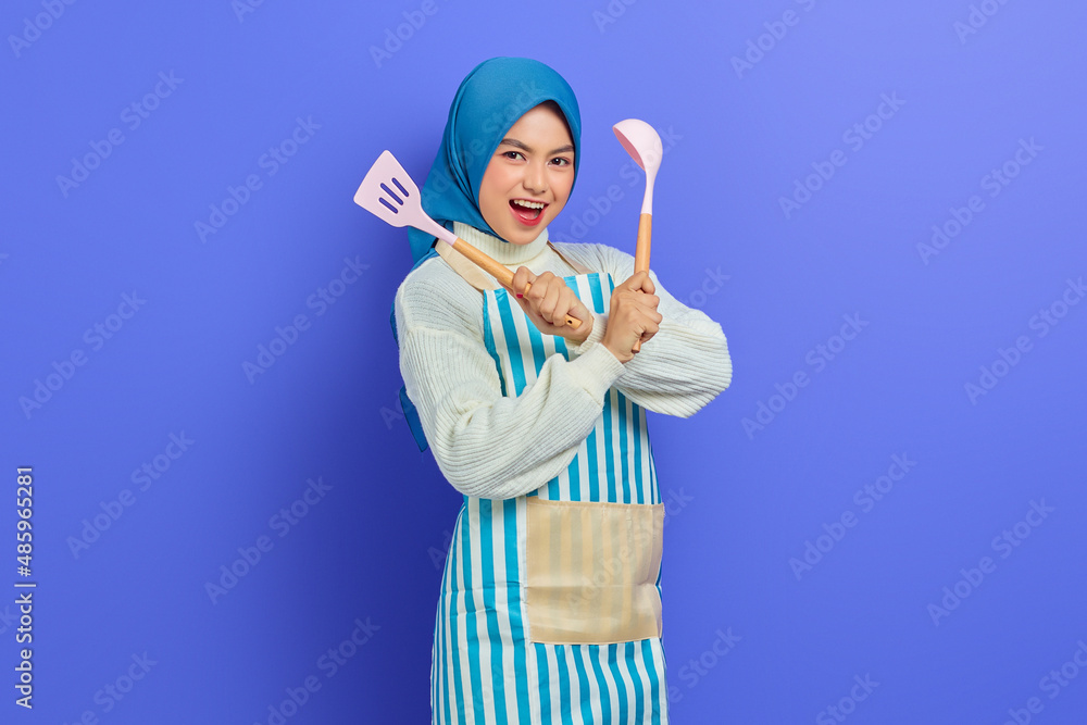 Excited beautiful Asian woman in white sweater covered in apron and hijab crossed hand hold spatula and spoon while doing housework isolated over purple background. Housekeeping concept Excited beautiful Asian woman in white sweater covered in apron and hijab crossed hand hold spatula and spoon while doing housework isolated over purple background. Housekeeping concept