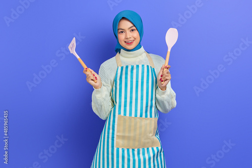 Cheerful beautiful Asian woman housewife in apron holding spatula and spoon soup while doing housework isolated over purple background. Housekeeping concept