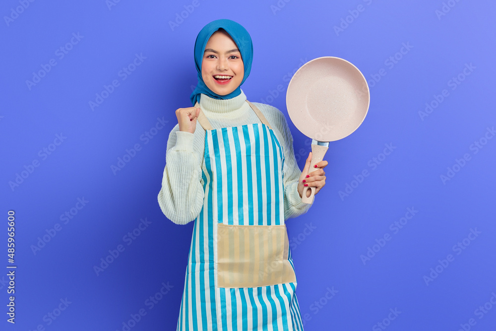 Portrait of excited young Asian Muslim woman wearing apron holding frying pan and celebrating luck isolated over purple background. Housekeeping concept Portrait of excited young Asian Muslim woman wearing apron holding frying pan and celebrating luck isolated over purple background. Housekeeping concept