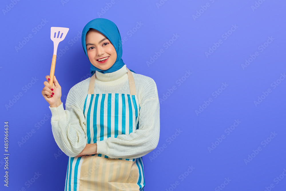 Cheerful beautiful Asian woman in white sweater covered in apron and hijab holding spatula, looking at camera doing housework isolated over purple background. Housekeeping concept Cheerful beautiful Asian woman in white sweater covered in apron and hijab holding spatula, looking at camera doing housework isolated over purple background. Housekeeping concept