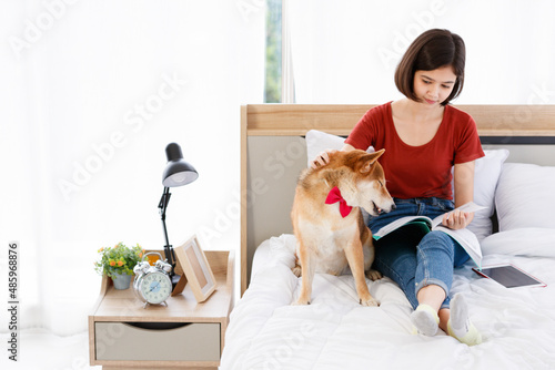 Little cute beautiful smart brown Japanese Shiba Inu dog wearing red bowtie sitting on bed together with Asian young female girl owner laying down leaning on pillow reading book and tablet in bedroom