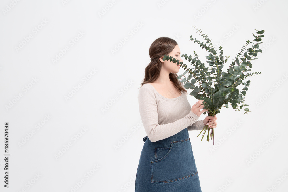 A horizontal photo. Pretty young girl florist in an apron holds a bouquet of eucalyptus in her hands and smiles. Girl small business entrepreneur. Prepares a large bouquet as a gift.