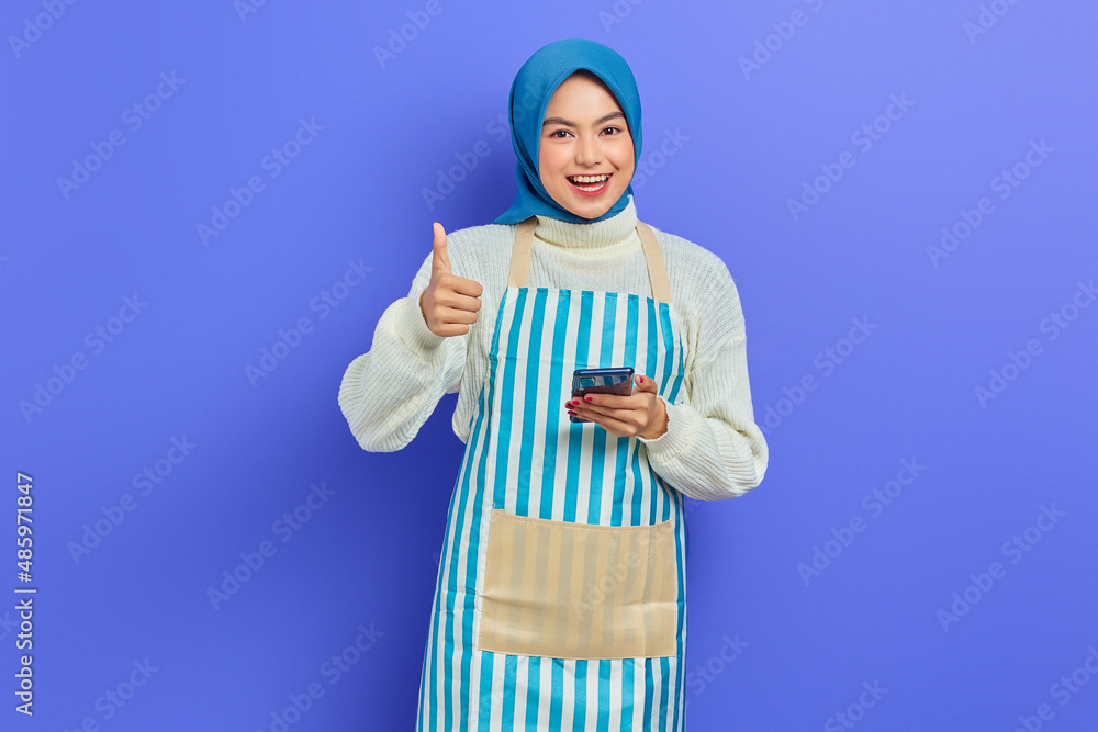 Portrait of smiling young Asian muslim woman in 20s wearing hijab and apron using mobile phone, showing thumb up gesture isolated over purple background. People housewife muslim lifestyle concept Portrait of smiling young Asian muslim woman in 20s wearing hijab and apron using mobile phone, showing thumb up gesture isolated over purple background. People housewife muslim lifestyle concept