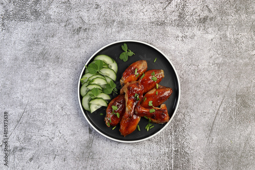 Honey Glazed Chicken Wings with sliced cucumbers and herbs on a round plate on a dark gray background. Top view, flat lay