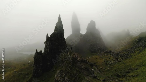 Man in dark clothes stands on top of a crag with in the background the enormously high rock peaks shrouded in fog called Old man of Storr rock in Skye mountains in Scotland. Drone dolly shot