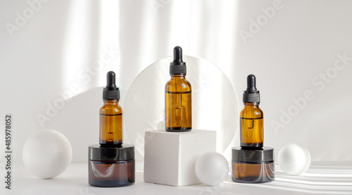 Composition of brown glass vials with dropper lids on geometric 3d podium. White background with rays of sunlight. Concept of packaging for cosmetic skin care products