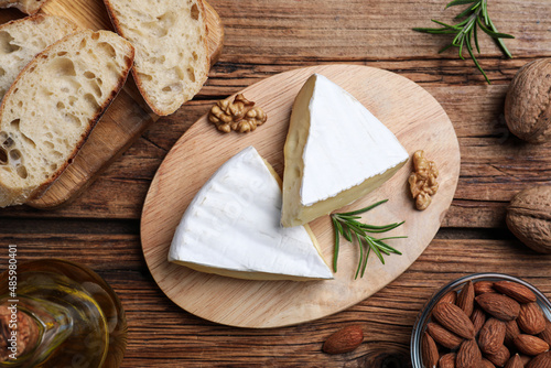 Tasty cut brie cheese with rosemary, bread and nuts on wooden table, flat lay