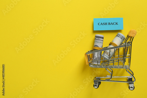 Card with word Cashback and rolled dollar banknotes in shopping cart on yellow background, flat lay. Space for text