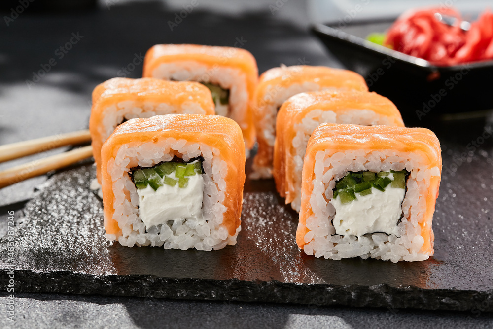 Maki sushi on dark stone table. Philadelphia maki with salmon. Sushi roll with cheese, cucumber inside, salmon outside. Style concept japanese menu with black background, leaves and hard shadow. Maki sushi on dark stone table. Philadelphia maki with salmon. Sushi roll with cheese, cucumber inside, salmon outside. Style concept japanese menu with black background, leaves and hard shadow.