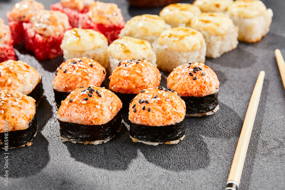 Hot maki set on dark slate. Baked sushi set with variety rolls. Sushi rolls with salmon,tobiko, crab and baked cheese topped. Style concept sushi menu with black background, leaves and hard shadow. Hot maki set on dark slate. Baked sushi set with variety rolls. Sushi rolls with salmon,tobiko, crab and baked cheese topped. Style concept sushi menu with black background, leaves and hard shadow.